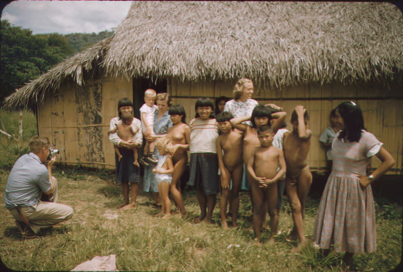 Marj Saint (holding son Philip), Elisabeth Elliot (head turned to the right), and Dayuma (far right end of group in red dress) with a group of Waorani women and children being filmed by unidentified photographer.