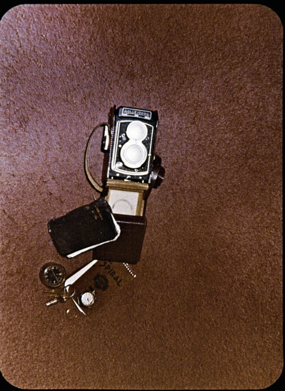 Image of Nate Saint's personal items recovered from Palm Beach. One frame from filmstrip <i>Unforgettable Friday</i>, created by Mission Aviation Fellowship in late 1956. Accompanying narration from Marjorie Saint for the frame: "The men of the ground party arrived back in Shell Mera and gave to each of us five wives the things they had found on the bodies of our husbands. I looked at Nate's camera, his airplane keys, the compass, his watch stopped at twelve minutes past three, then his tiny notebook used as a diary during those days and the little black New Testament and Psalms."