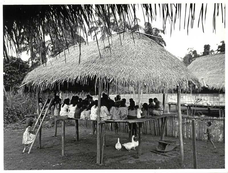 Black and white photograph of Waorani gathered in thatched structure. The reverse of the photograph includes two lengthy captions. The fist typed caption reads: "The Auca Church designed and built by themselves with a palm-thatched roof and bamboo floor. Here Dr. Edman held the first Bible Conference for these new Christians, and for the first time they celebrated the Lord's Supper. For bread they used boiled yuca (manioc or arrowroot) and for the wine they used banana drink in a gourd brought from the forest." The second handwritten caption reads: "Church designed and built by the Aucas, Tiwaeno. Bible class taught here Nov 4-5, 1965 by VRE. Mark 4 - Parable of the Sower (+ recd [?] from Ball Seed Co.). [?] meeting, Thurs. 11/4, by moonlight. Friday, 11/5, [?] on Baptism, Mark 16. 11 baptized, beginning with Dabo, and then Kimo and Dyuwi. Then the Lord's supper - first time for the Aucas - Mark 14 + 1 Cor 11, boiled [?] and banana drink. New song led by Dyuwi. The commissioning of Dyuwi and Tonia - Read Acts 13 - laying on of hand w/ Geo. M. Traber[?], Kimo, and VRE. Church lies between Rachel's and Dayuma's (background). Aucas planned church is for expansion when their [?] come to the Lord. Sept. '65."