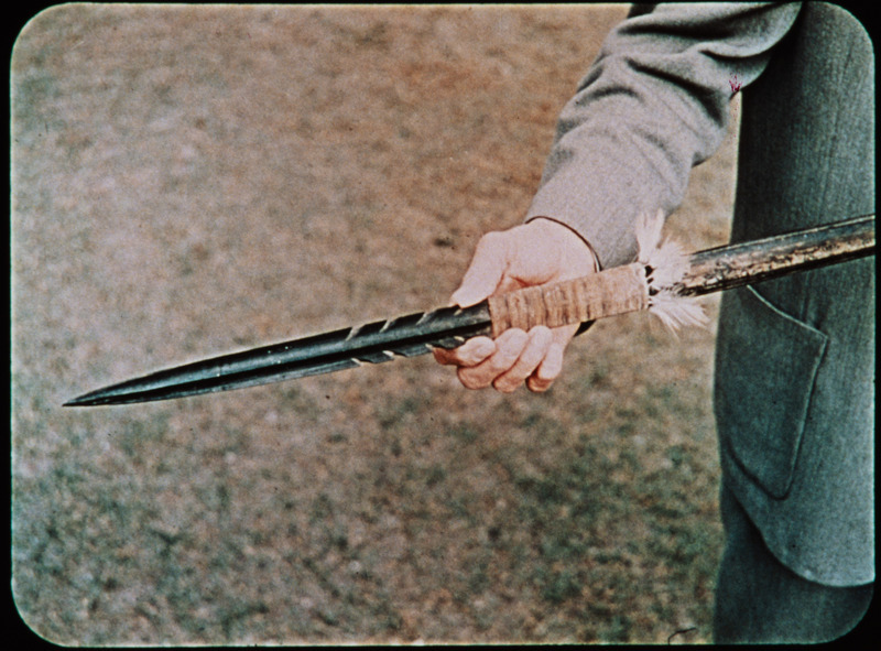 Image of unidentified man holding a wooden spear recovered from Palm Beach. Frame from <i>Mid-Century Martyrs</i> filmstrip developed by HCJB, Mission Aviation Fellowship, and the five widows in 1956. Along with the earlier frame which pictured Marj Saint and Steve Saint at the radio in Shell Mera, the accompanying audio or transcript read: "Five days later the rescue squad located their bodies that had been pierced by the long Auca spears on the shore of the Curaray River, deep in Auca territory."