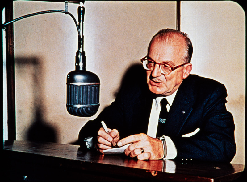 Color image of radio missionary and HCJB co-founder Clarence Jones at microphone in HCJB radio station, Quito, Ecuador. Jones reported the public first announcement of the missing missionaries over HCJB on Monday night, January 9, 1956.  Frame from <i>Mid-Century Martyrs</i> filmstrip developed by HCJB, Mission Aviation Fellowship, and the five widows in 1956.