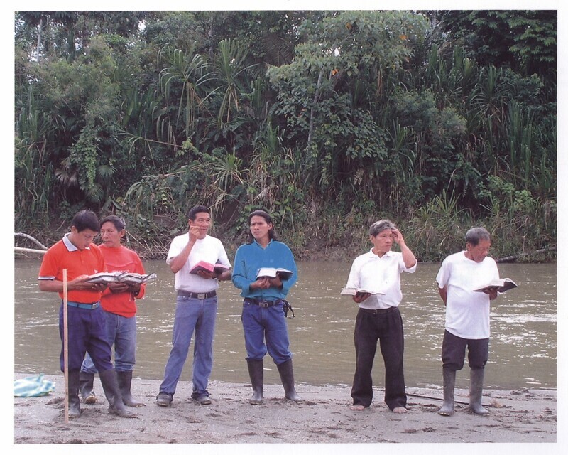 Color photograph of Waorani Christians leading memorial and baptism service near "Palm Beach" on the 50th anniversary of the death of the five missionaries. Paa (an elder in the Waorani church) is pictured with his hand on his head, holding Wao New Testament. Others are unidentified. Photograph taken during Ken Fleming's 2006 anniversary visit to Ecuador. A former missionary to Africa, Ken Fleming was the brother of Peter Fleming.