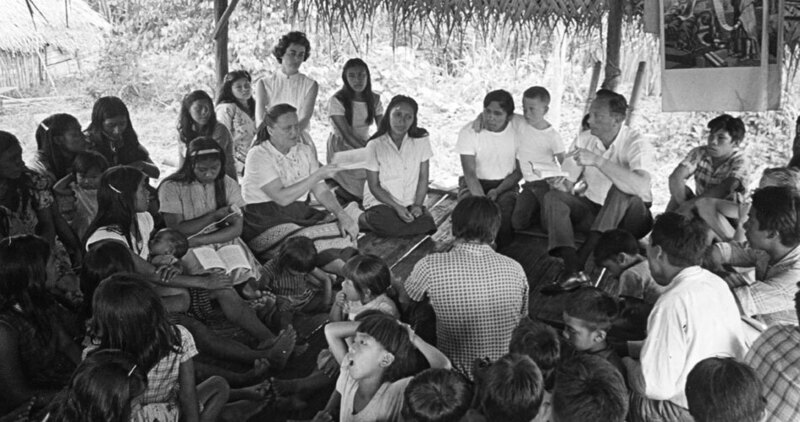 Rachel Saint with Dayuma and Waorani Indians in Tewæno community. Caption written on back, likely by Rachel Saint: “Oucaye, sent to us in a miraculous way from the down-river group, read her own copy—being able to half-read and half-memorize her Easter verse. Many times she has expressed her joy at having 'God’s carving' for herself, a wonderful preparation for the trip home to tell the others as God works it out." 