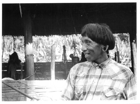 Photograph of Geuquita in western clothing in front of home. Caption from V. Raymond Edman: "Gikita, the oldest of the Aucas, and his house high on the hill across the river from the Auca Village. He is the historian of the tribe with many memories of what the ancients taught and did." September 1965.