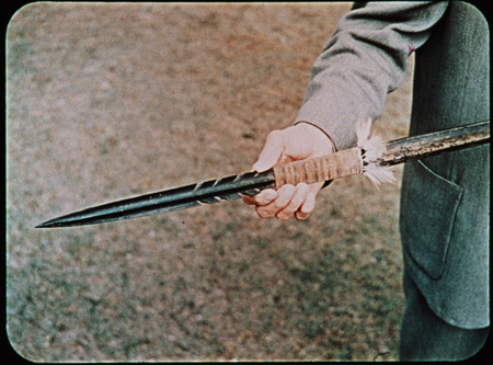 Image of unidentified man holding a wooden spear recovered from Palm Beach. Frame from <i>Mid-Century Martyrs</i> filmstrip developed by HCJB, Mission Aviation Fellowship, and the five widows in 1956. Along with the earlier frame which pictured Marj Saint and Steve Saint at the radio in Shell Mera, the accompanying audio or transcript read: "Five days later the rescue squad located their bodies that had been pierced by the long Auca spears on the shore of the Curaray River, deep in Auca territory."