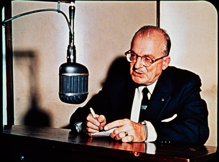 Color image of radio missionary and HCJB co-founder Clarence Jones at microphone in HCJB radio station, Quito, Ecuador. Jones reported the public first announcement of the missing missionaries over HCJB on Monday night, January 9, 1956.  Frame from <i>Mid-Century Martyrs</i> filmstrip developed by HCJB, Mission Aviation Fellowship, and the five widows in 1956.