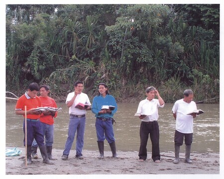 Color photograph of Waorani Christians leading memorial and baptism service near "Palm Beach" on the 50th anniversary of the death of the five missionaries. Paa (an elder in the Waorani church) is pictured with his hand on his head, holding Wao New Testament. Others are unidentified. Photograph taken during Ken Fleming's 2006 anniversary visit to Ecuador. A former missionary to Africa, Ken Fleming was the brother of Peter Fleming.