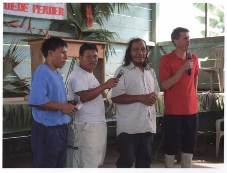Color photograph of Quemo (Kimo) and Brethren missionary Dan Rogers (in red) leading service of Waorani church at Toñampade. Although church buildings have been established at Toñampade and Tewæno, many Waorani Christians practice in home churches based around the traditional Waorani kinship community. Photograph taken during Ken Fleming's 2006 anniversary visit to Ecuador. A former missionary to Africa, Ken Fleming was the brother of Peter Fleming.